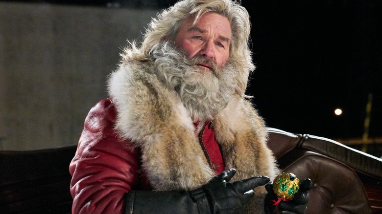 White-bearded man in a red fur-trimmed coat holds a small gold ornament while seated outdoors at night.