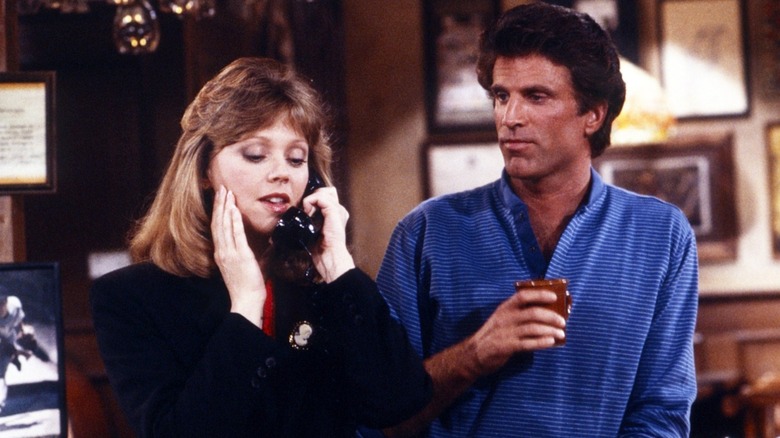 A woman speaks on a phone while a man holding a drink stands beside her indoors.