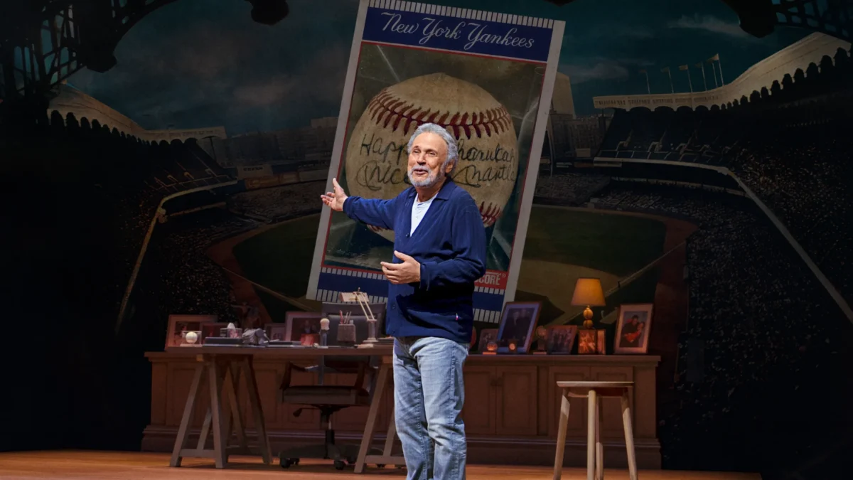 An older man gestures onstage beside a desk, stool, and large New York Yankees baseball card backdrop.