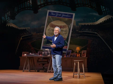 An older man gestures onstage beside a desk, stool, and large New York Yankees baseball card backdrop.