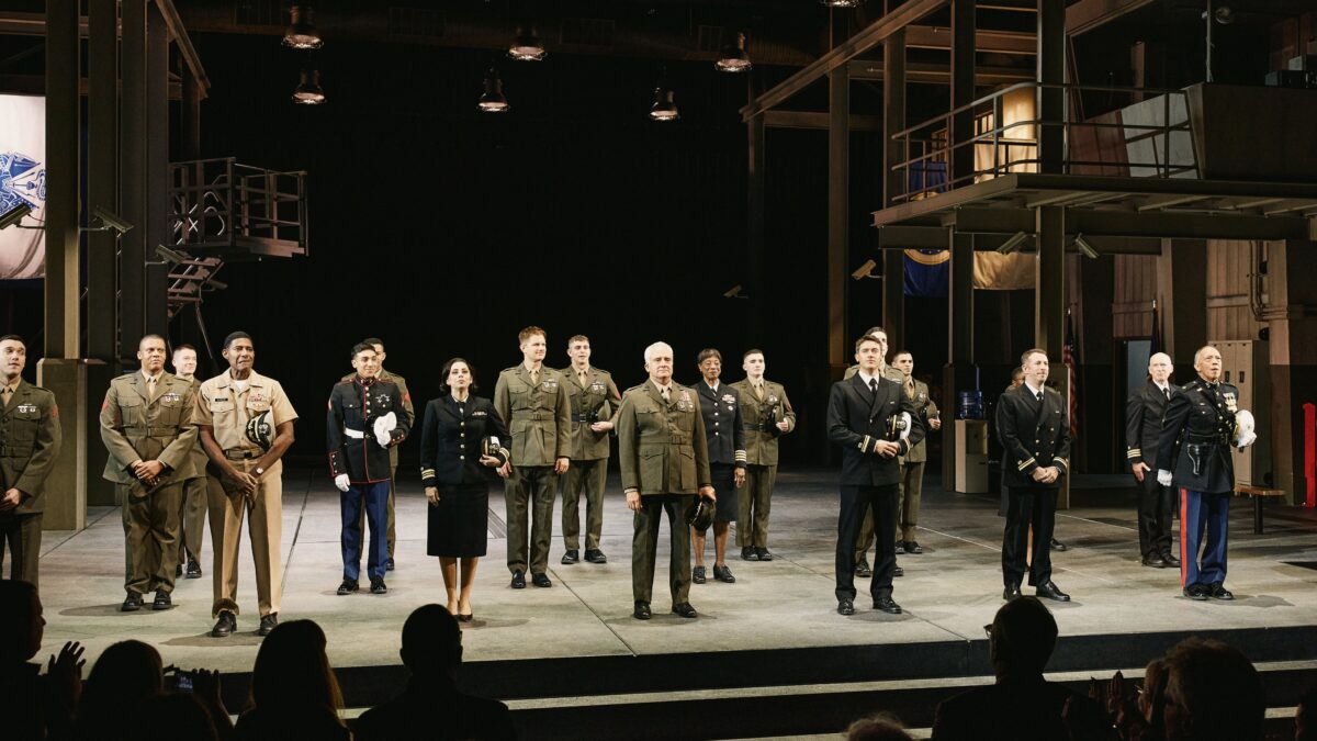 People in military uniforms stand across a stage before an applauding audience.