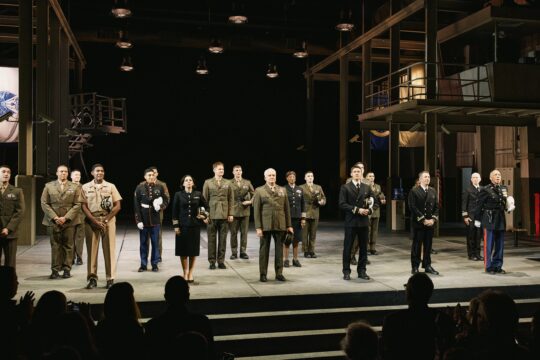 People in military uniforms stand across a stage before an applauding audience.
