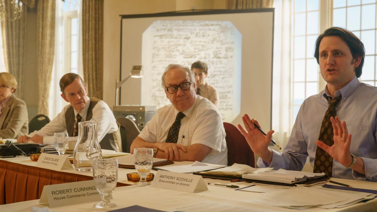 Several people sit at a conference table with papers, water glasses, nameplates, and a projected document behind them.