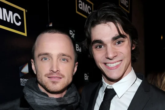 Two men pose closely at a black AMC-branded backdrop, one smiling broadly in a suit and tie.
