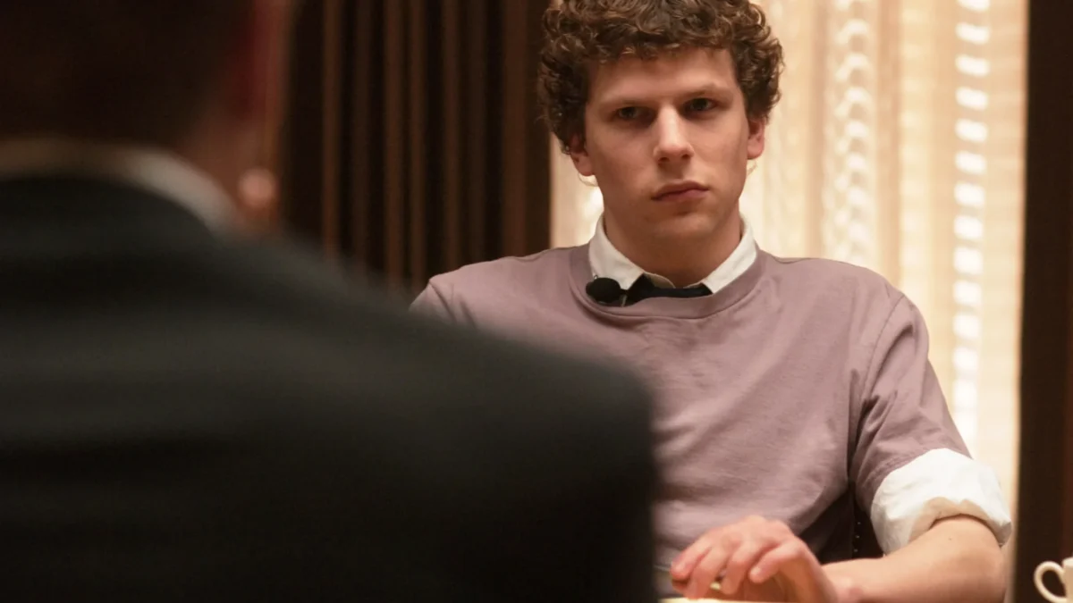 A curly-haired man in a lavender shirt sits at a table, facing a blurred figure in the foreground.