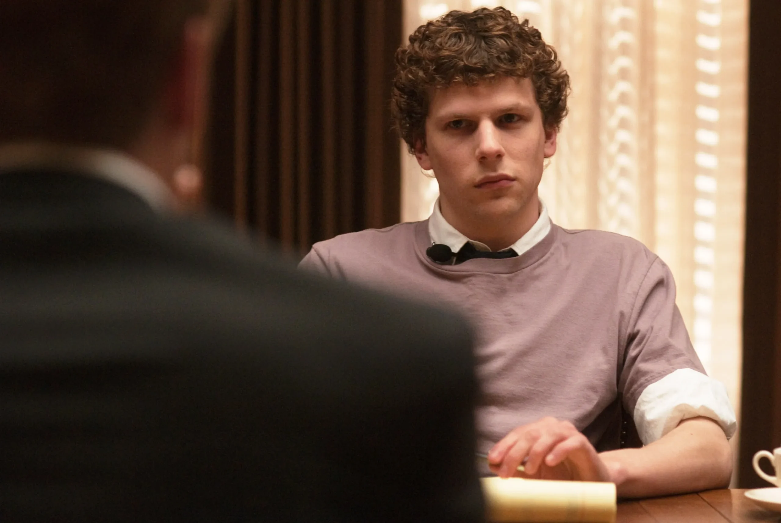 A curly-haired man in a lavender shirt sits at a table, facing a blurred figure in the foreground.