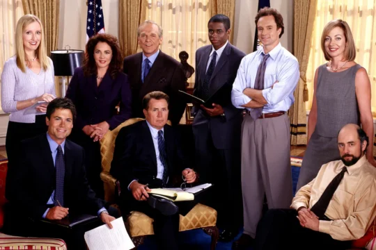 Nine formally dressed adults pose together in a stately room with flags, curtains, papers, and notebooks.