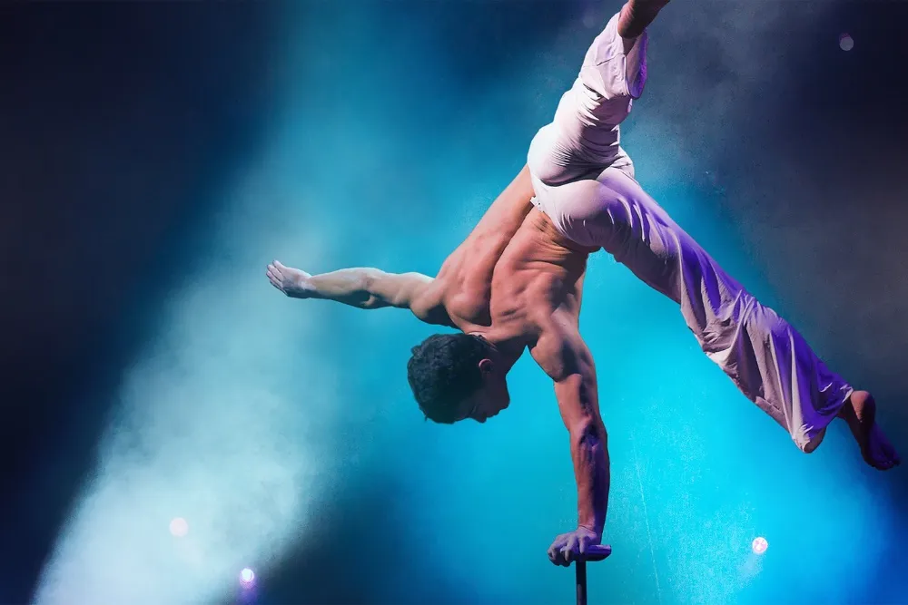 A shirtless performer balances upside down on one hand against blue stage lights and haze.