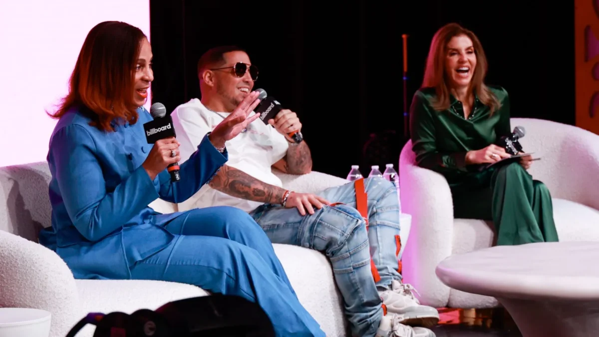 Three panelists sit on white chairs, holding branded microphones during a brightly lit stage discussion.