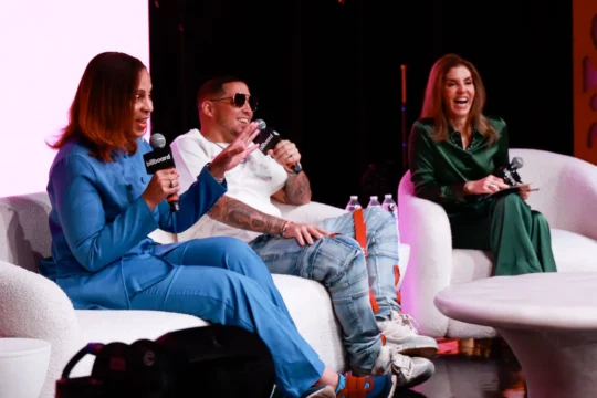 Three panelists sit on white chairs, holding branded microphones during a brightly lit stage discussion.