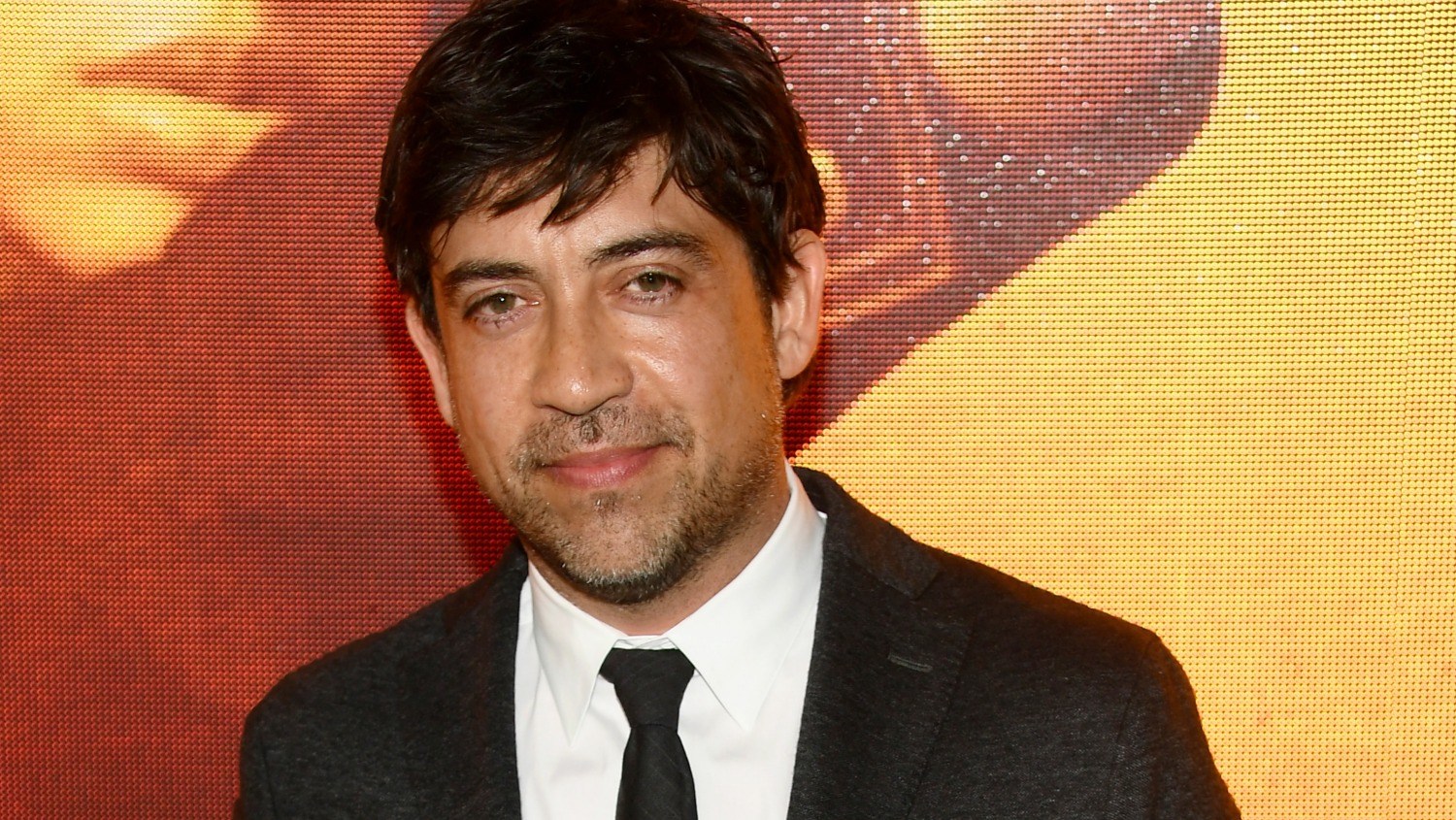 A man in a dark suit and black tie stands before an orange patterned backdrop.