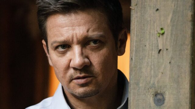 A serious-looking man peers from behind a wooden post in dim light.
