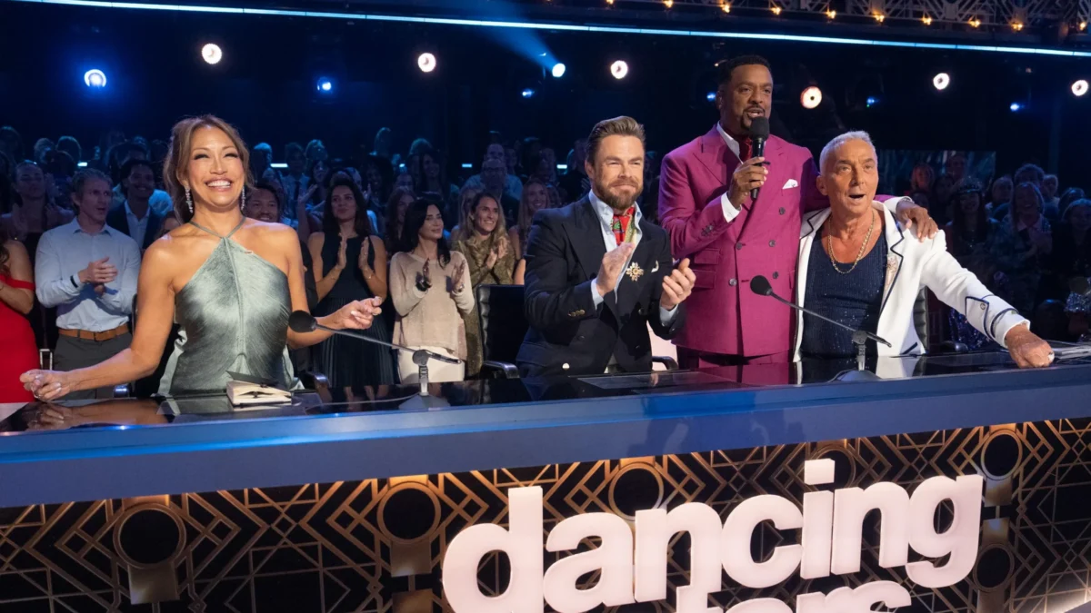 Four people at a judges’ desk react with smiles, applause, and a microphone before a clapping audience.