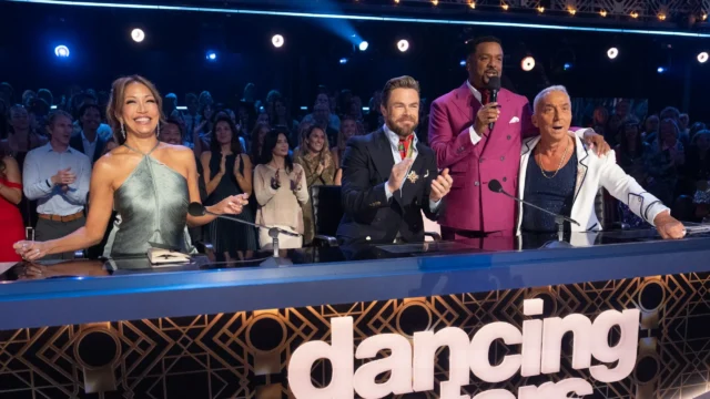 Four people at a judges’ desk react with smiles, applause, and a microphone before a clapping audience.