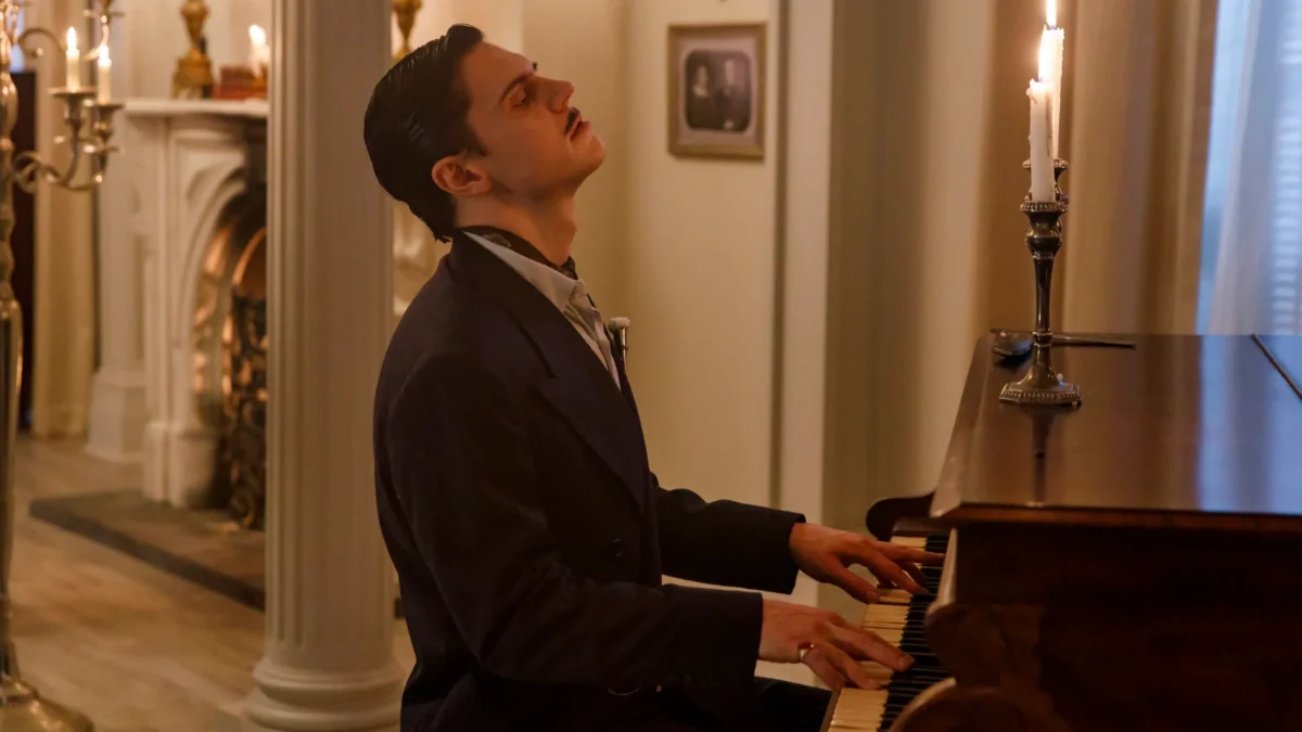Mustached man in a suit plays piano by candlelight in an ornate room.