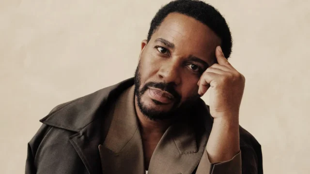 A man with short hair and a beard rests his head on his hand against a beige background.