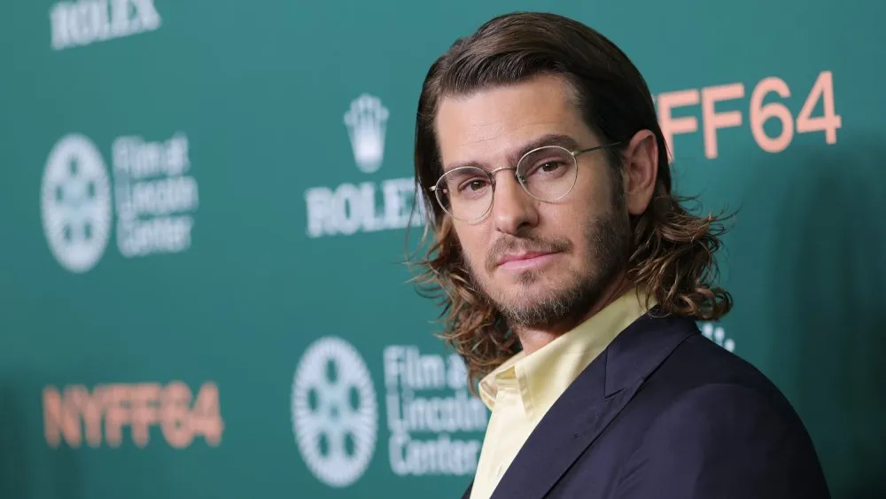 A man with shoulder-length hair and glasses poses in a dark suit before a green event backdrop.