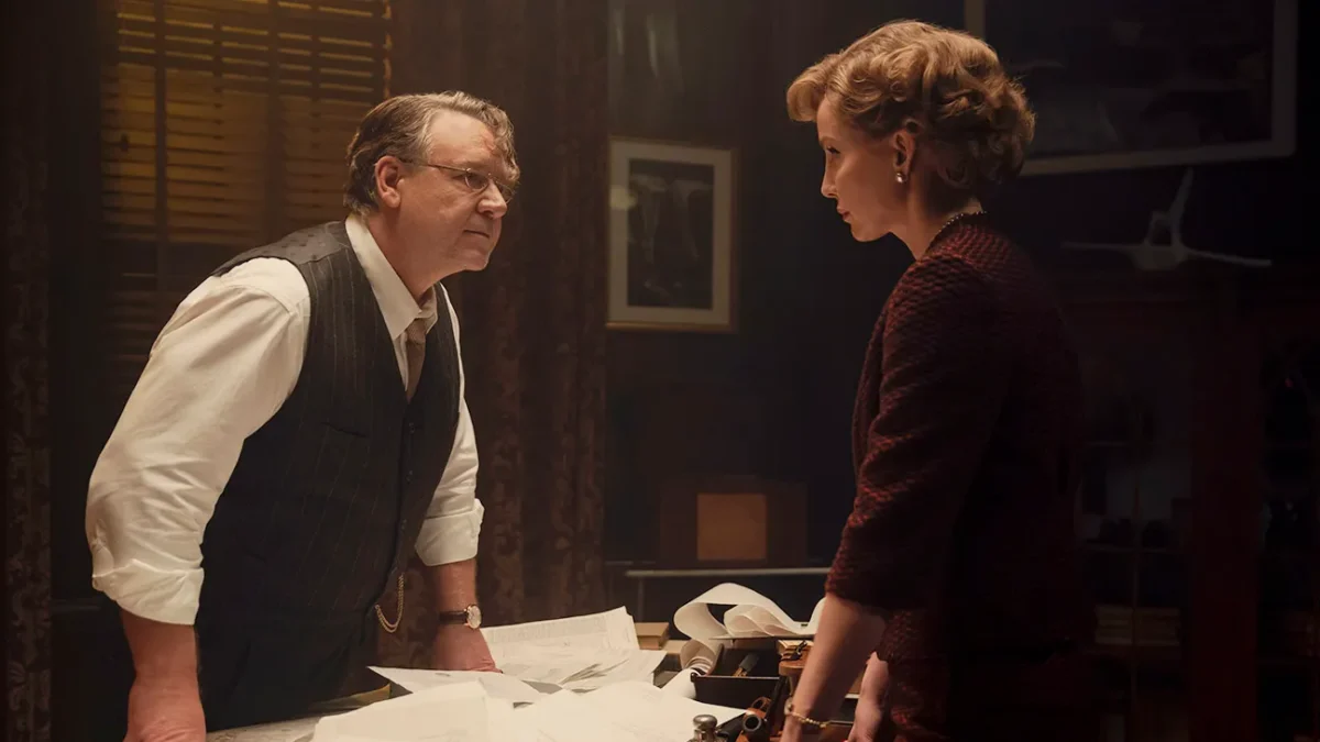 "A man and woman in formal attire have a serious discussion in a dimly lit office, standing over a desk covered with papers and old-fashioned equipment, creating a tense atmosphere."
