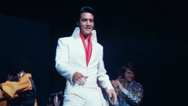 A performer in a white suit and red shirt stands on a dark stage with others behind him.