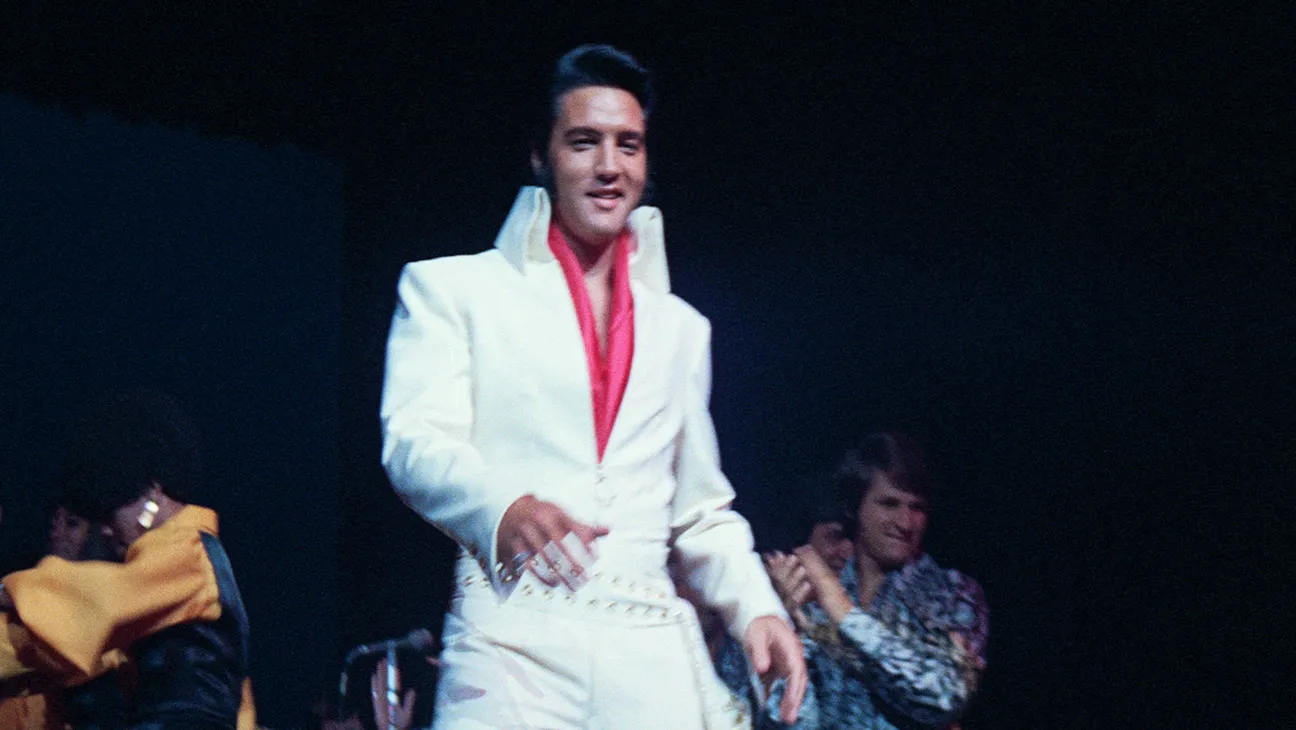 A performer in a white suit and red shirt stands on a dark stage with others behind him.