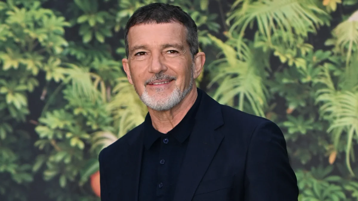 A man with a salt-and-pepper beard wearing a black suit stands before a leafy green backdrop.