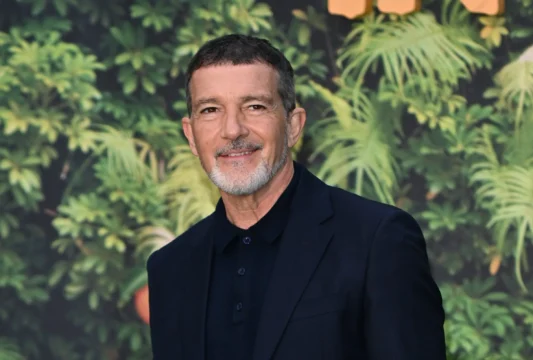 A man with a salt-and-pepper beard wearing a black suit stands before a leafy green backdrop.