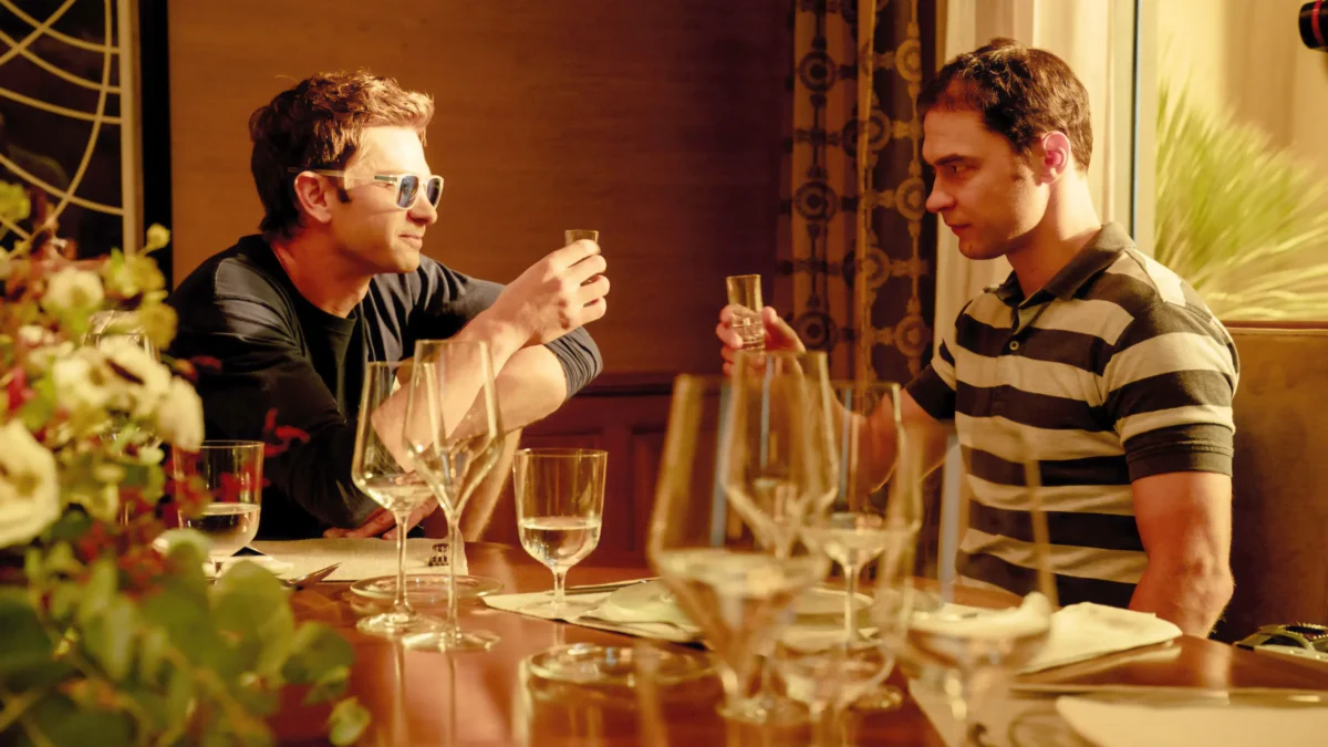 Two men seated at a dining table raise small glasses amid wine glasses and warm lighting.