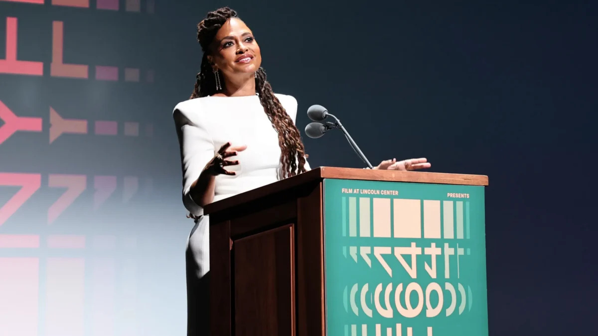A woman in a white dress gestures behind a podium with microphones on a dark stage.