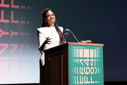 A woman in a white dress gestures behind a podium with microphones on a dark stage.