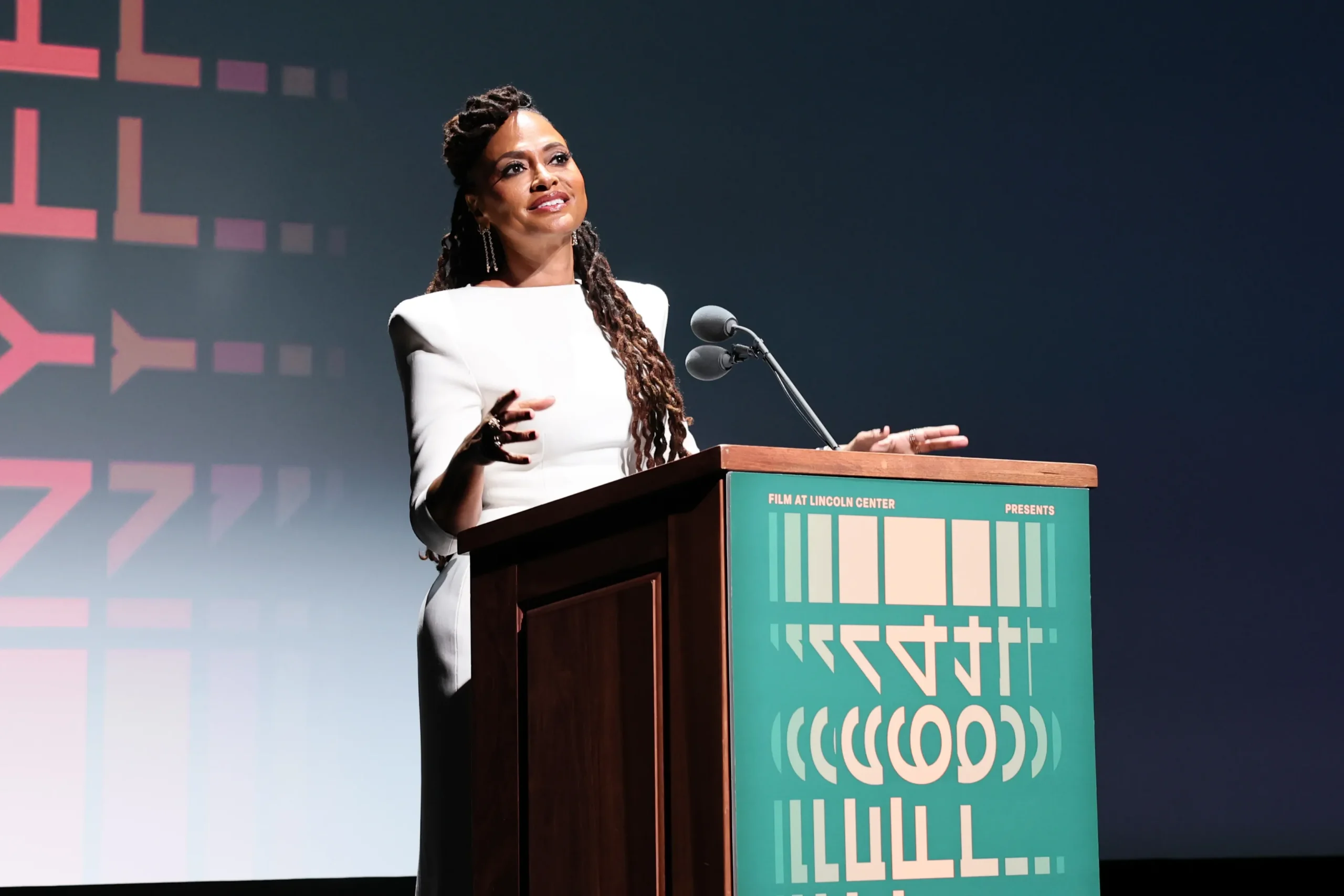 A woman in a white dress gestures behind a podium with microphones on a dark stage.