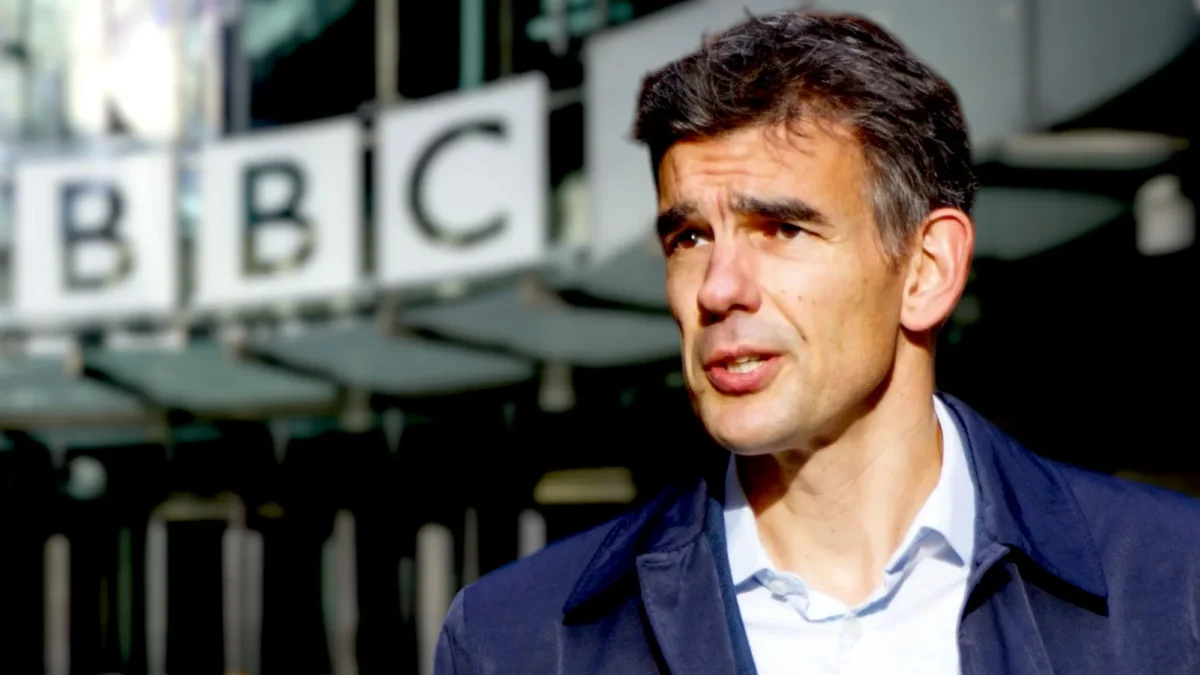 A man in a blue jacket stands outdoors with a blurred BBC sign in the background.