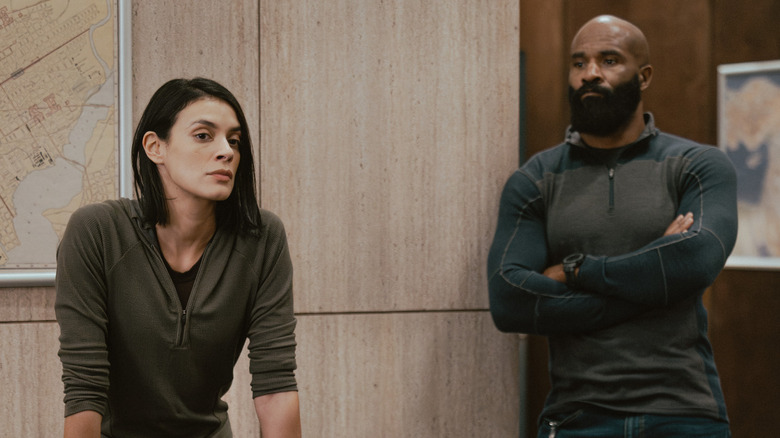 A woman leans forward in a room while a bearded man stands behind her with arms crossed.