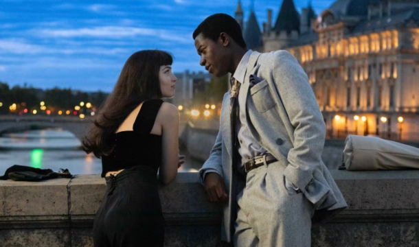 A woman and man lean on a riverside wall at dusk, facing each other with illuminated buildings behind them.