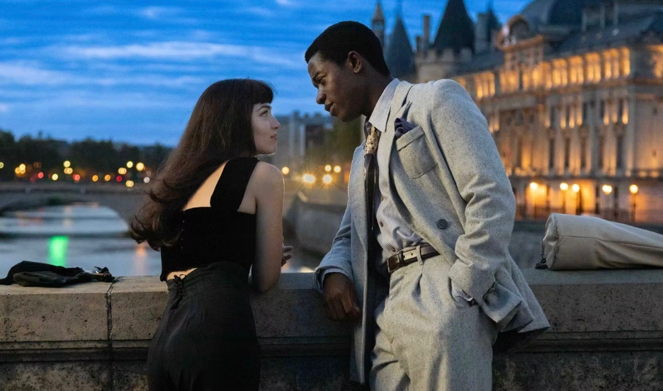 A woman and man lean on a riverside wall at dusk, facing each other with illuminated buildings behind them.