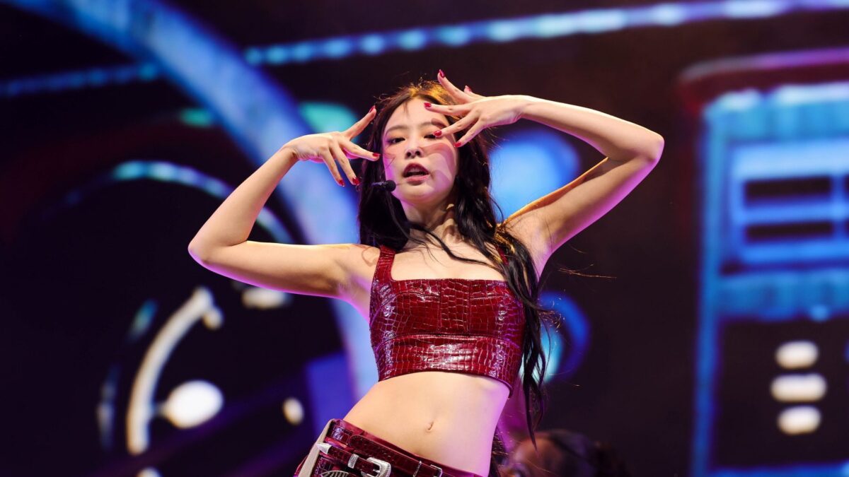 A performer in a red outfit poses with hands framing her face on a brightly lit stage.