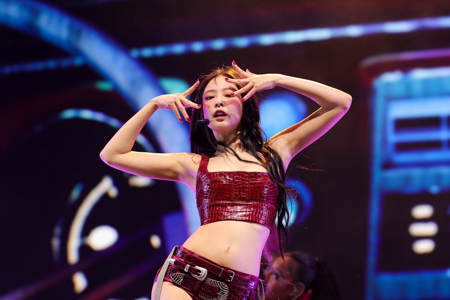 A performer in a red outfit poses with hands framing her face on a brightly lit stage.