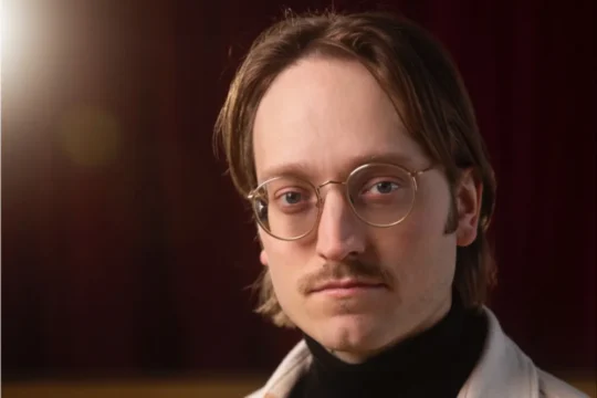 Close-up portrait of a man with round glasses and a mustache against a dark, blurred background.