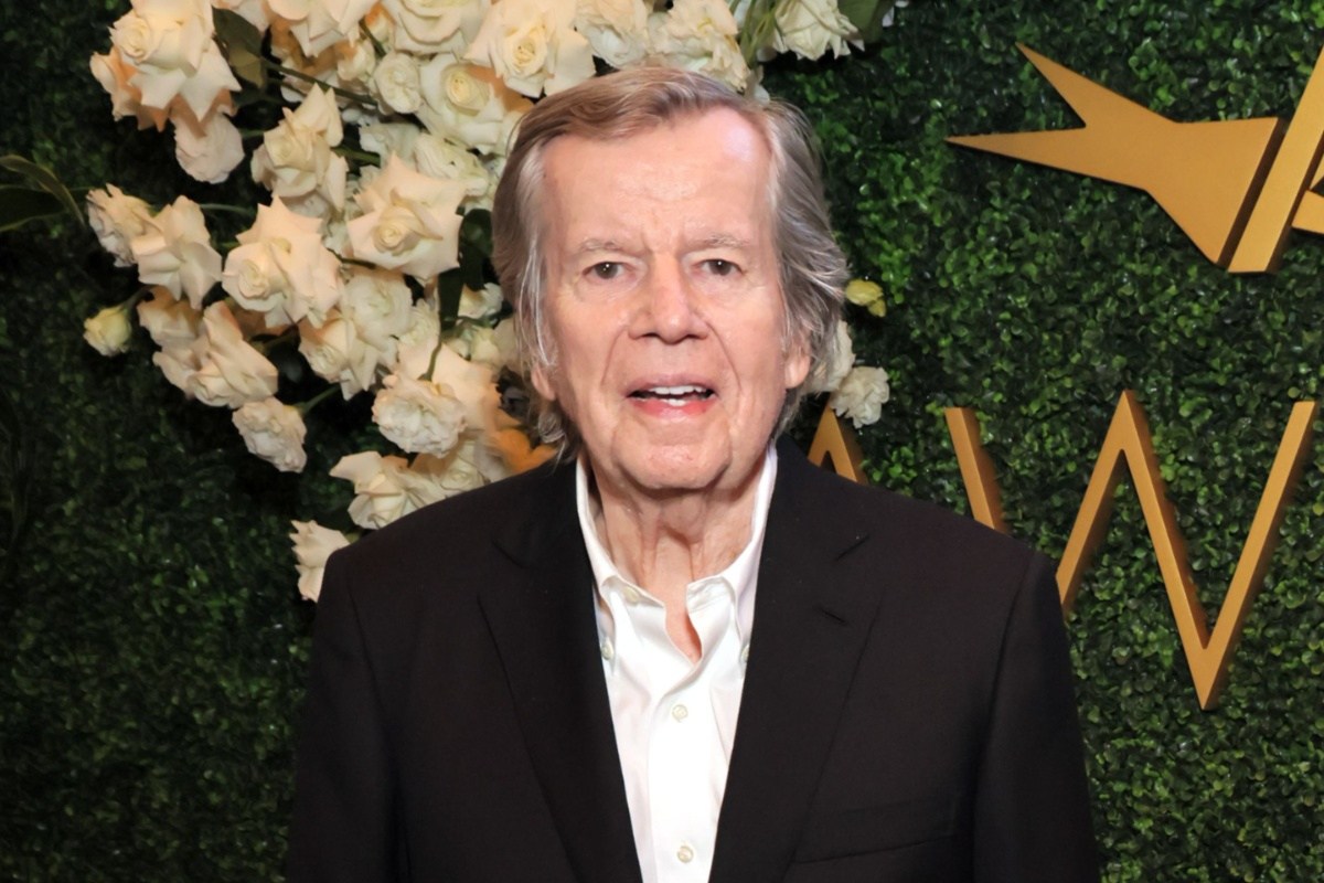 An older man in a black blazer stands before white roses and a green backdrop with gold shapes.