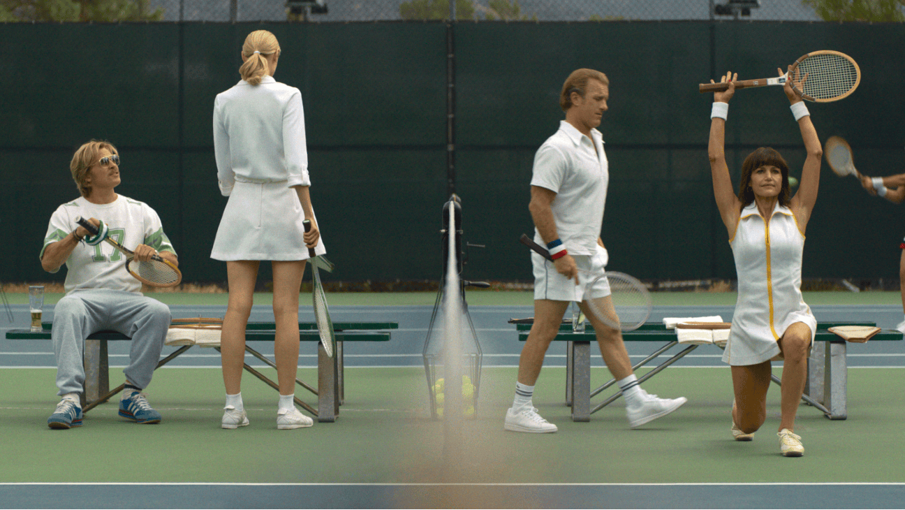 Four tennis players in white outfits are on a court, one kneeling and holding a racket overhead.