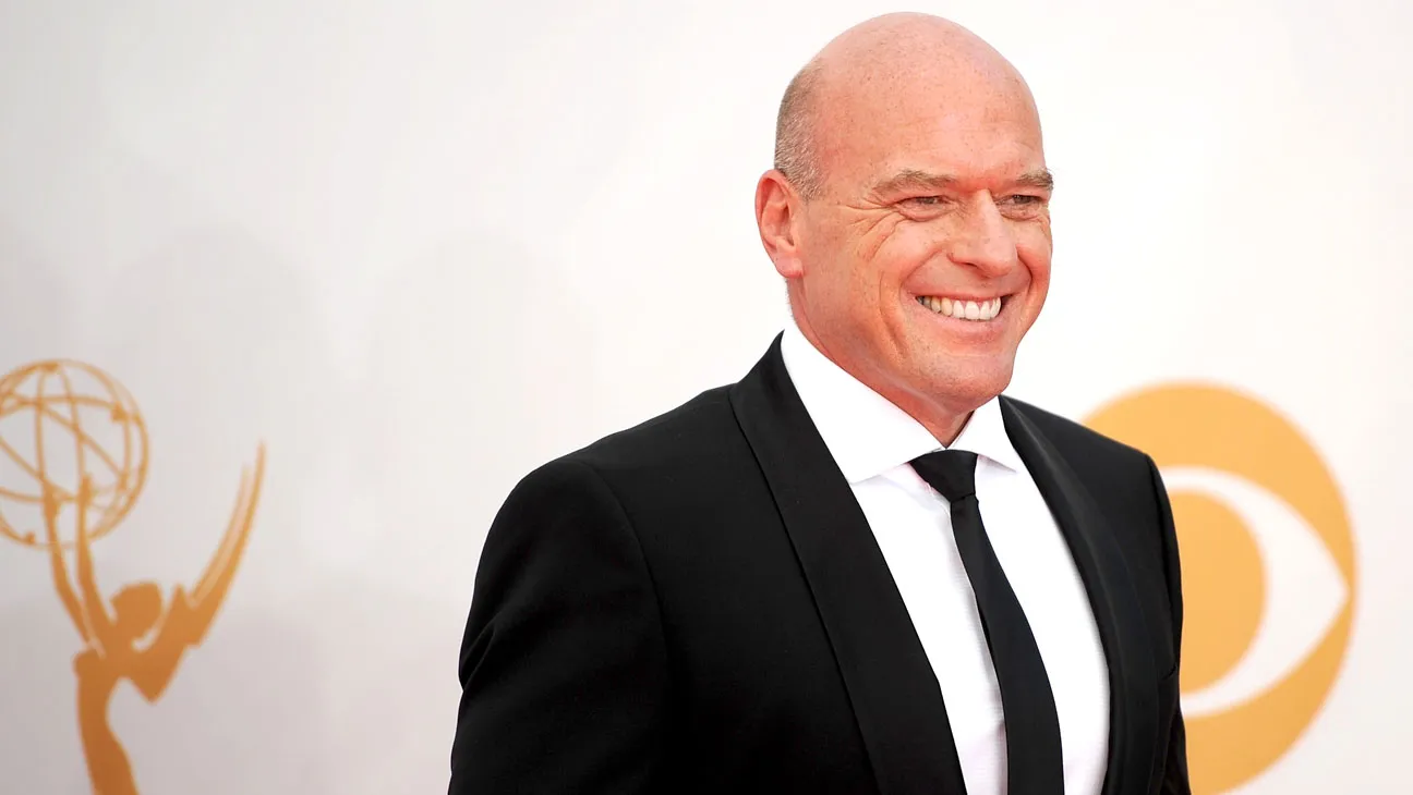 A bald man in a black suit smiles in front of a white backdrop with gold logos.