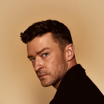 A man with short dark hair and stubble looks over his shoulder against a beige background.