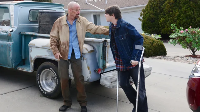 A bald man speaks to a young man using crutches beside a weathered blue pickup truck in a driveway.