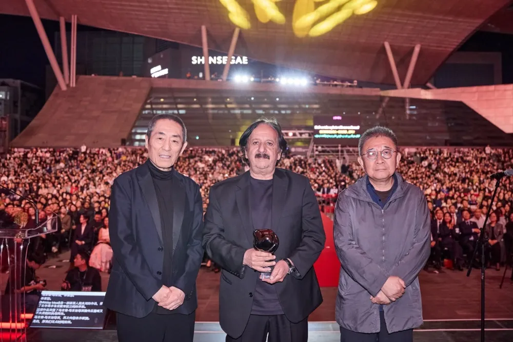 Three men stand on a stage before a large nighttime crowd, with one holding a trophy.