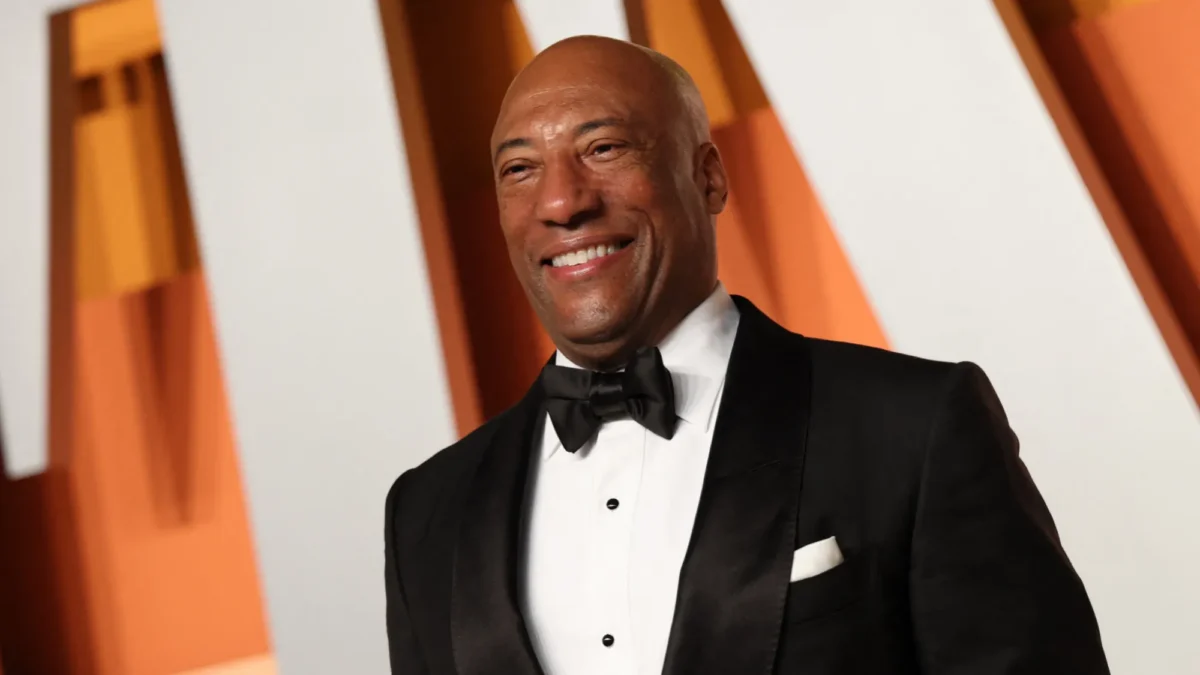 A smiling bald man in a black tuxedo stands before orange and white vertical panels.