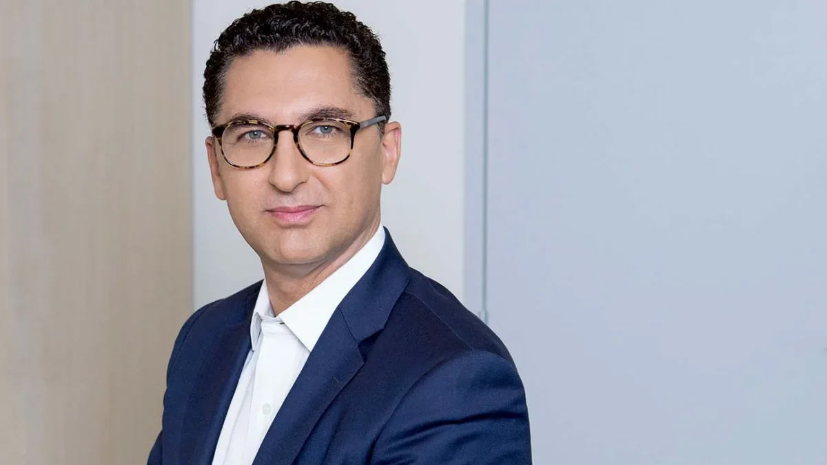 Man in glasses wearing a navy suit jacket and white shirt poses against a light wall.