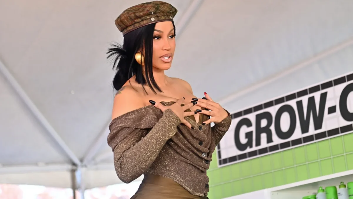 A woman in a brown outfit and cap poses with hands near her chest beside a green display.