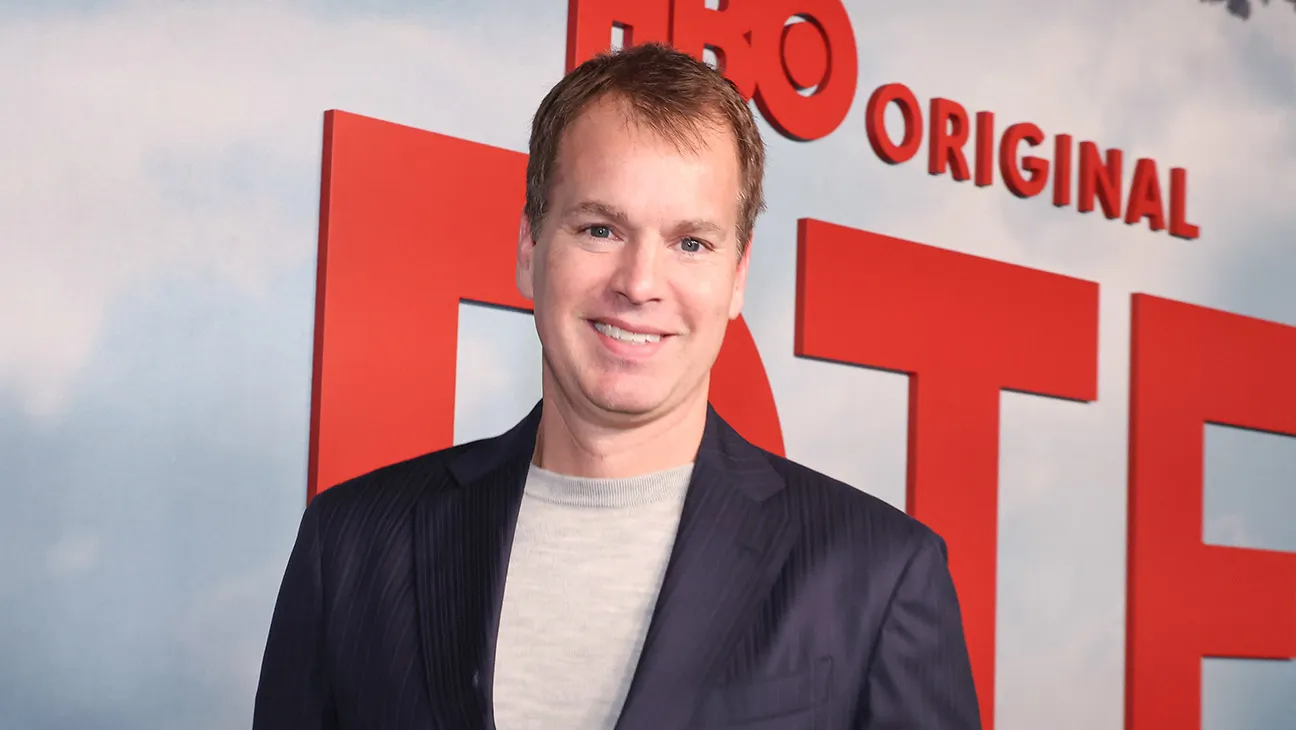 A smiling man in a dark blazer stands in front of large red HBO Original signage.