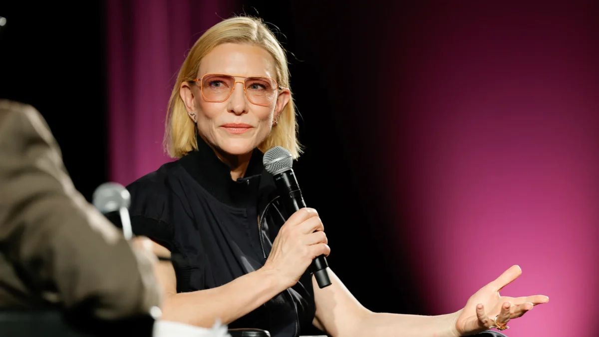 A blonde woman wearing orange glasses holds a microphone while speaking onstage against a pink backdrop.