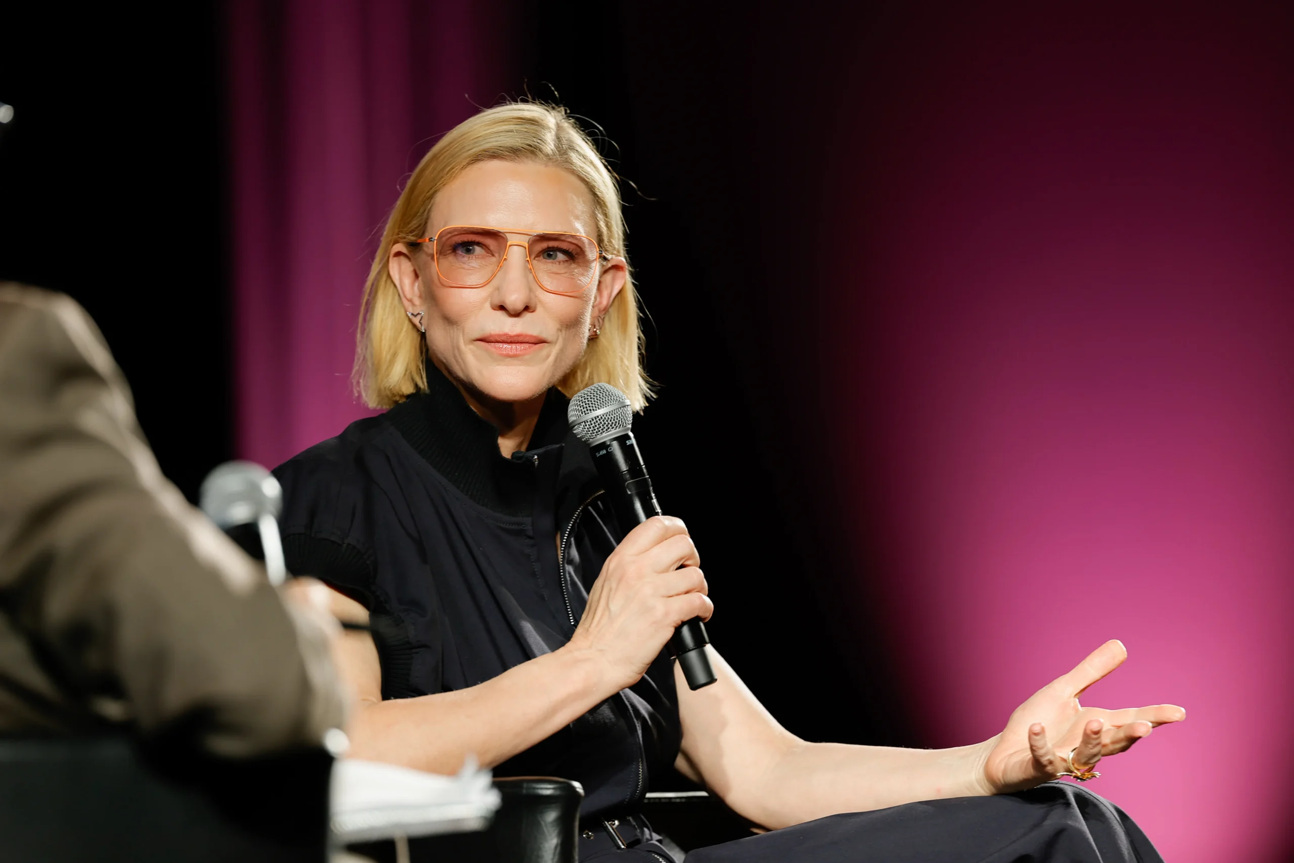 A blonde woman wearing orange glasses holds a microphone while speaking onstage against a pink backdrop.