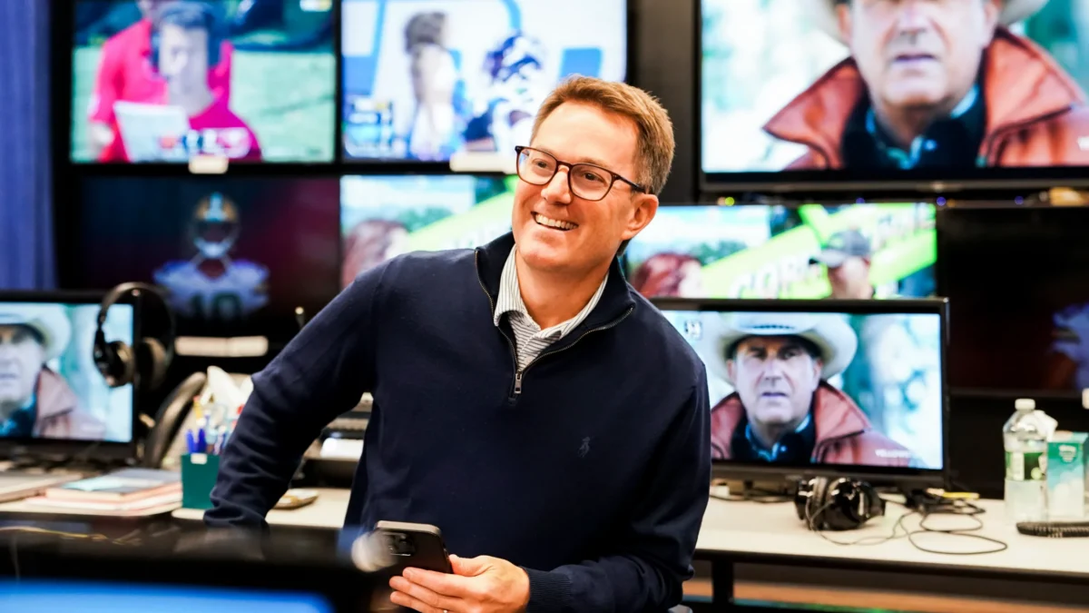 A smiling man with glasses holds a phone in a control room filled with television monitors.